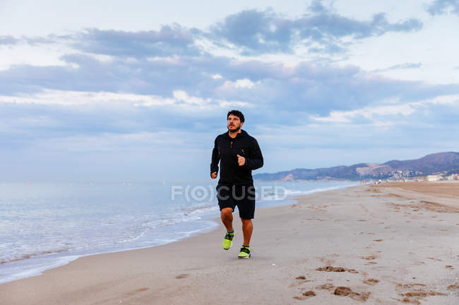 Чоловік у спортивному одязі бігає на піску в морі під час тренування на відкритому повітрі на пляжі на заході сонця — стокове фото