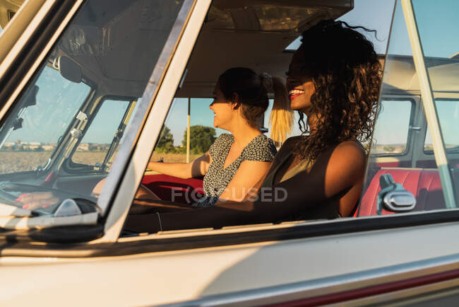 Vista lateral de dos mujeres jóvenes sentadas dentro de una furgoneta vintage y sonriendo mientras viajan en la naturaleza juntas - foto de stock