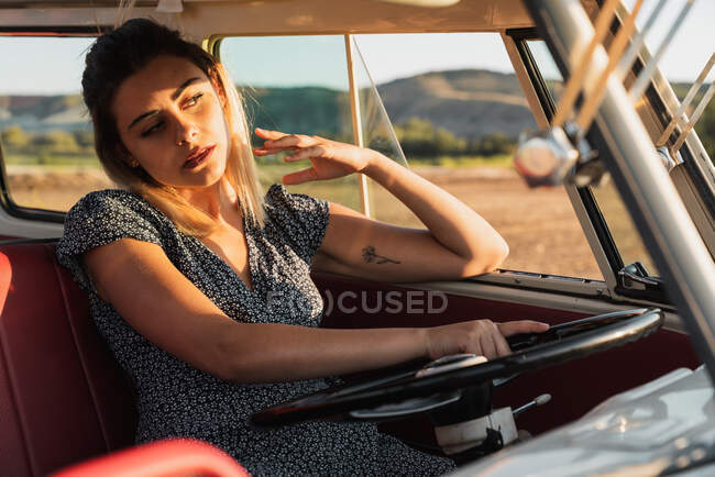 Young female sitting on driver seat and steering vintage van while traveling in countryside on sunny day — Stock Photo
