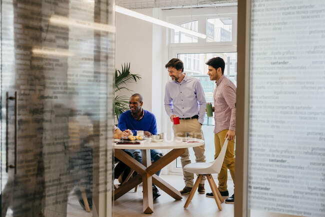 Alegre grupo de hombres multiétnicos sentados y de pie a la mesa en la oficina. - foto de stock