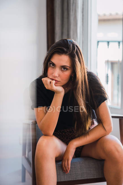 Retrato de mujer morena joven en camiseta negra y pantalones cortos sentados en sillón en la habitación sobre fondo de ventana y cortina - foto de stock
