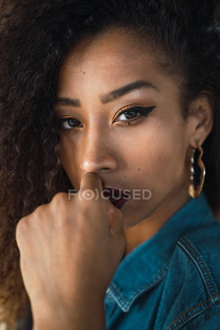 Jeune femme afro-américaine avec un maquillage lumineux et des cheveux bouclés foncés en chemise en denim regardant la caméra — Photo de stock