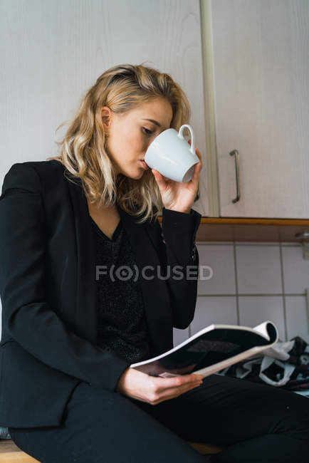 Випадкова розслаблена жінка має каву, сидячи на кухонній стійці і читаючи книгу — стокове фото