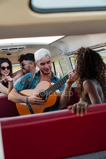Grupo de jóvenes riendo y escuchando chico guapo tocando la guitarra acústica dentro de la camioneta vintage en la naturaleza - foto de stock