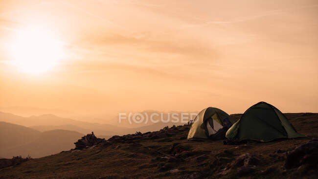 Personas anónimas sentadas cerca de dos tiendas de campaña en la cima de la colina en el fondo del majestuoso cielo matutino con sol naciente - foto de stock