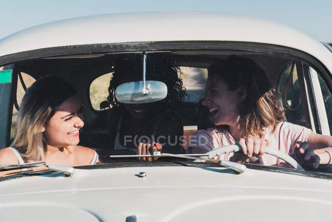 Group of happy multiethnic women in car driving together in bright sunlight and laughing — Stock Photo