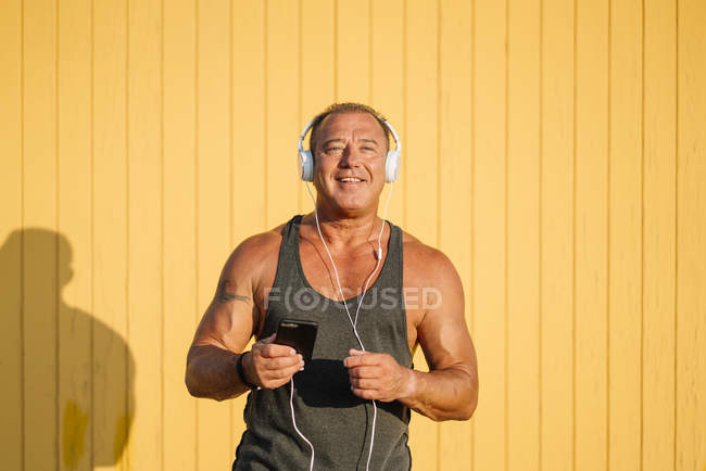 Homem mais velho forte posa com fones de ouvido no fundo amarelo — Fotografia de Stock