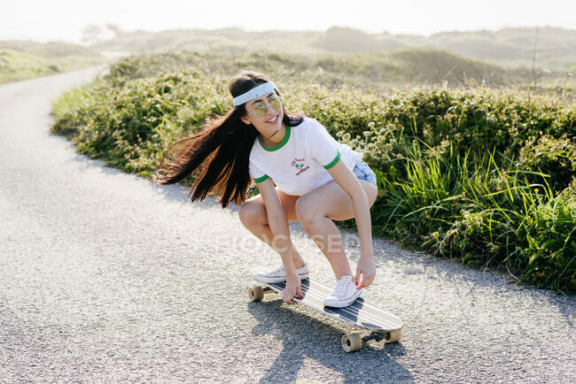 Menina casual alegre em óculos de sol com cabelo acenando montando placa longa na estrada pavimentada na natureza. — Fotografia de Stock