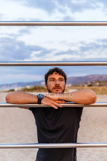 Hombre barbudo reflexivo en ropa deportiva apoyado en la escalera y mirando hacia otro lado mientras descansa durante el entrenamiento al aire libre en la playa al atardecer - foto de stock