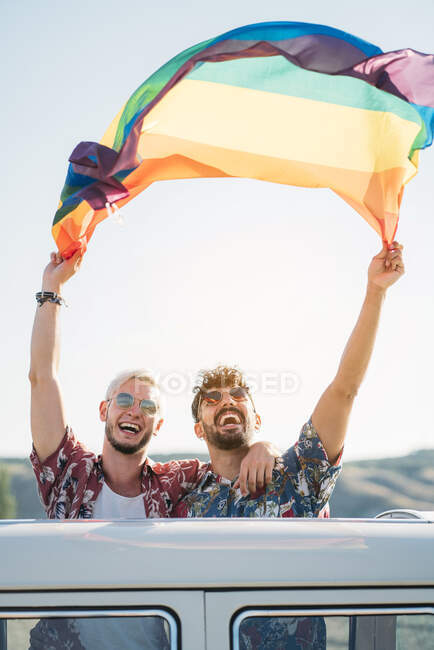 Zwei junge Männer umarmen sich und schwenken LGBT-Flagge über den Köpfen, während sie in einem Retro-Van mit geöffnetem Dach in der Natur stehen — Stockfoto