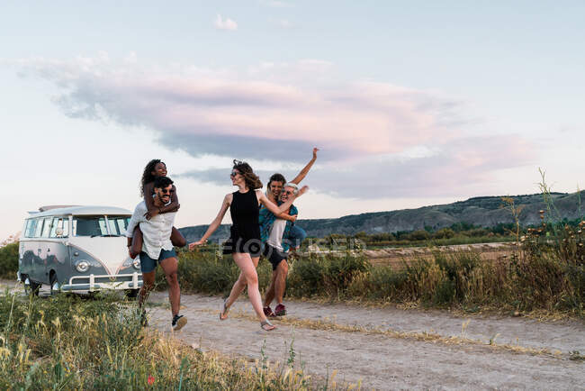 Two men giving piggyback ride to women and running along countryside road while spending time in nature together — Stock Photo