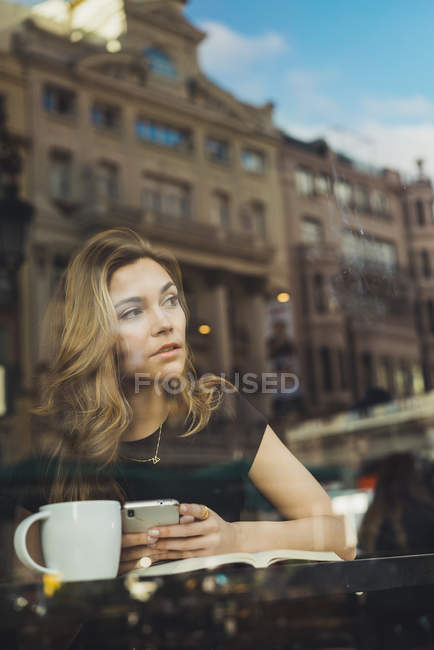 Young woman with smartphone, coffee cup and book sitting in cafe behind window — Stock Photo