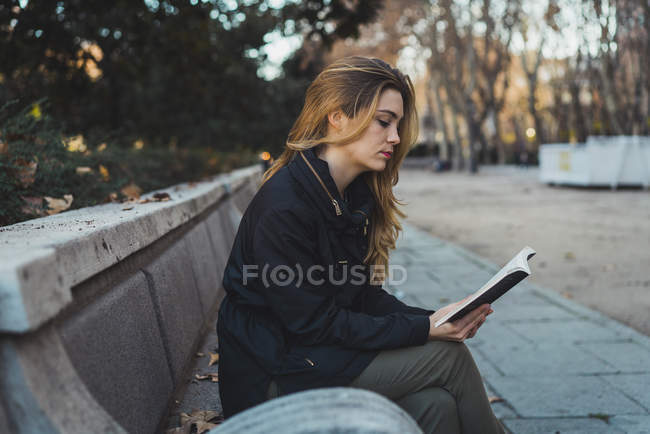 Young woman sitting on park bench and reading book — Stock Photo