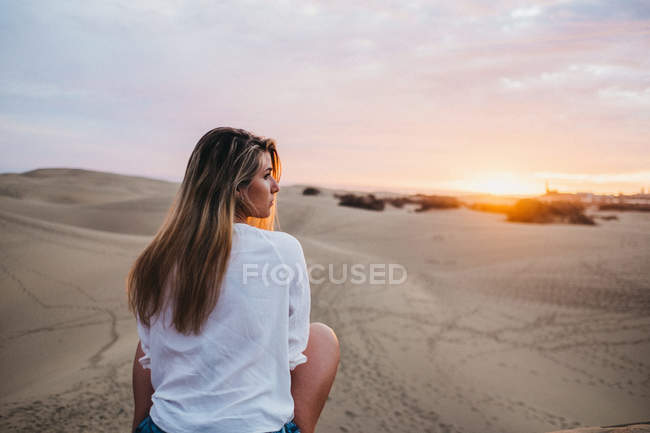 Young woman in white t-shirt sitting on sand at sunset and looking over shoulder — Stock Photo