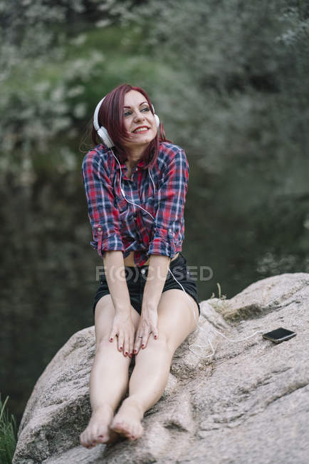 Рыжая девушка слушает музыку у реки — стоковое фото
