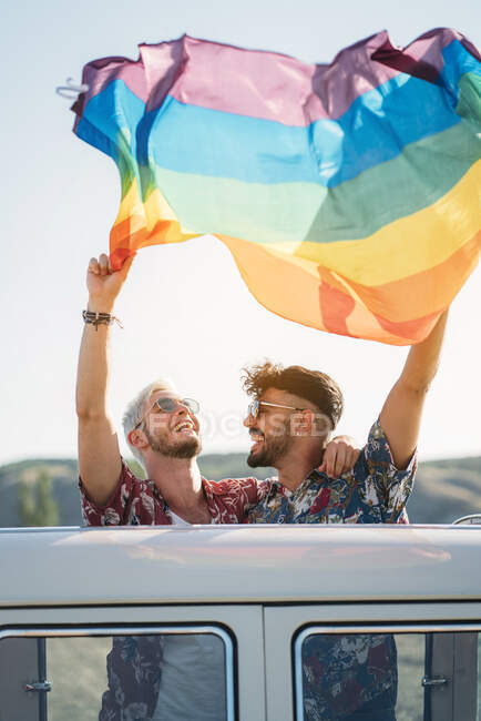 Dois jovens abraçando e segurando a bandeira LGBT acenando sobre as cabeças enquanto? de pé dentro da van retro com telhado aberto na natureza — Fotografia de Stock