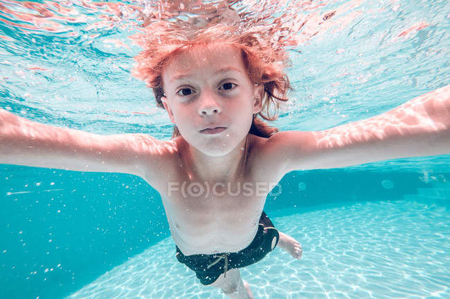 Рыжеволосый ребенок ныряет в воду и смотрит в камеру на фоне прозрачной воды — стоковое фото