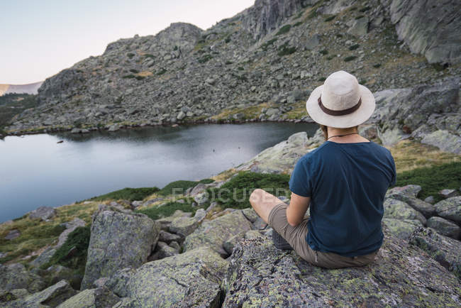Joven con sombrero sentado en las rocas cerca del lago y mirando a la vista - foto de stock