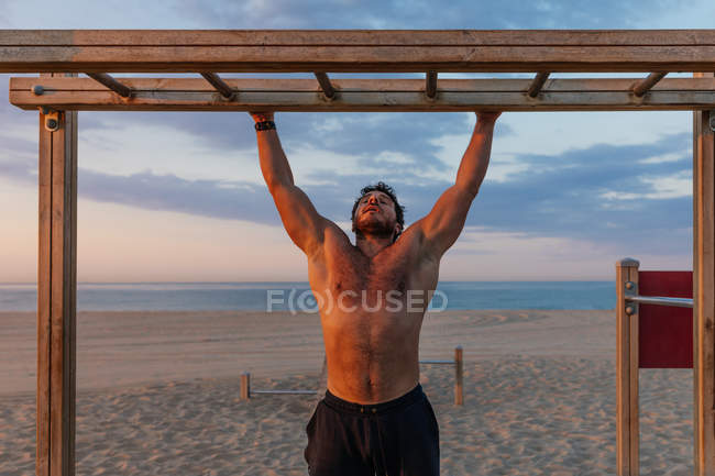 Бородатый мужчина без рубашки поднимается по деревянной лестнице во время тренировки на пляже — стоковое фото