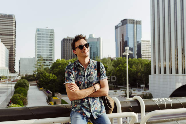 Fröhlicher junger Mann mit Sonnenbrille lehnt am Geländer und schaut in der Stadt weg — Stockfoto