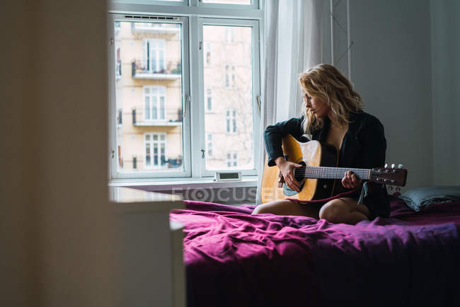 Блондинка молода жінка грає на гітарі на ліжку вдома — стокове фото