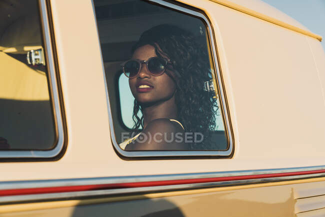 Beautiful African-American woman in sunglasses looking out window while traveling inside retro van on sunny day — Stock Photo