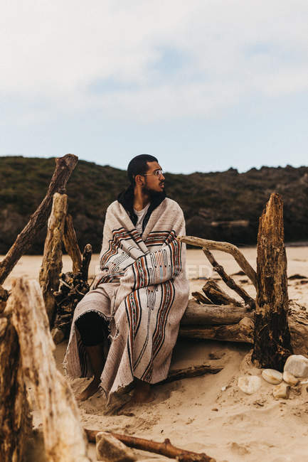 Bonito homem envolto em cobertor ornamental e olhando para longe enquanto sentado em armadilhas na costa arenosa — Fotografia de Stock
