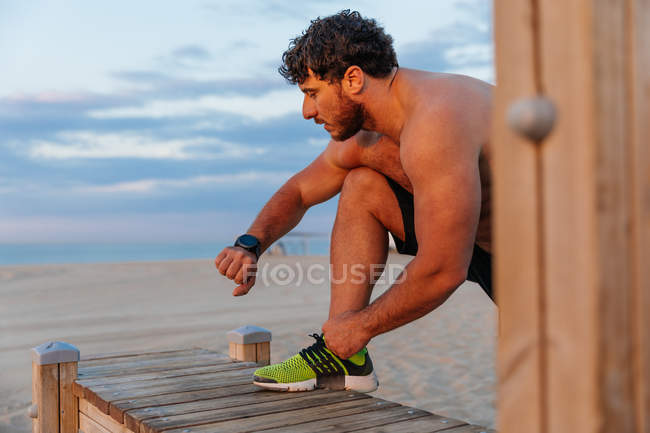 М'язистий бородатий чоловік перевіряє сучасний фітнес-трекер під час тренування на пляжі — стокове фото
