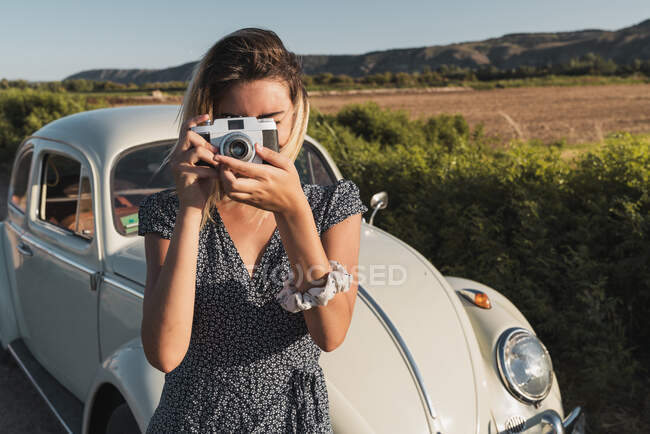 Woman in dress taking photo with retro camera standing on background of green summer landscape in sunlight — Stock Photo