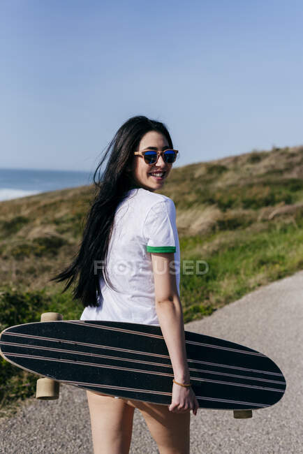 Back view of cheerful trendy brunette holding skating board and walking on rural road in sunlight. — Stock Photo