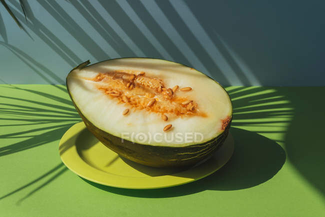 Halbe Melone auf Teller auf blauem und grünem Hintergrund mit Schatten von Palmblättern — Stockfoto