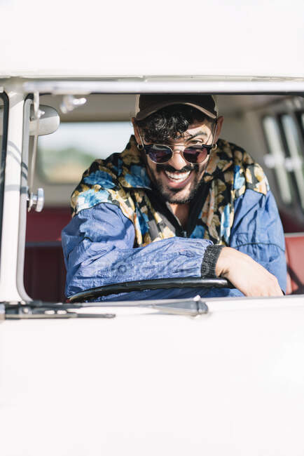 Привабливий молодий чоловік у стильному одязі, який спирається на кермо і дивиться на камеру, сидячи в старовинному фургоні під час подорожі по природі. — Stock Photo