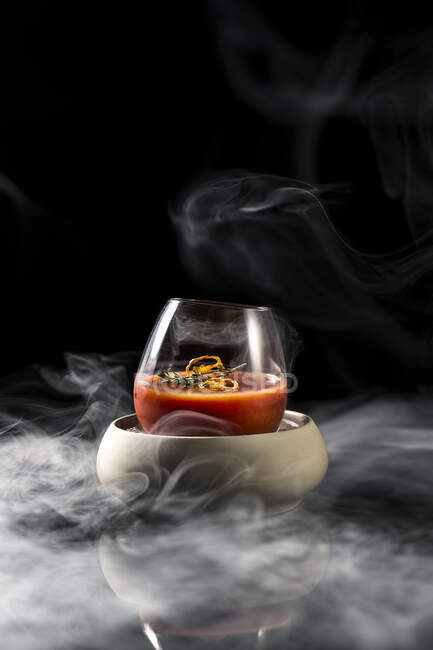 Zusammensetzung des Glases in einer Schüssel mit rotem, würzigem Alkoholcocktail gefüllt und auf dem Tisch in Rauch vor schwarzem Hintergrund serviert — Stockfoto