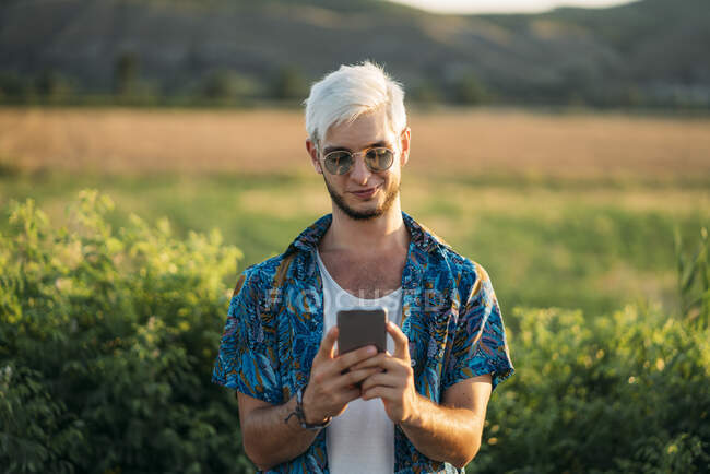 Beau jeune homme en tenue élégante riant et naviguant smartphone tout en se tenant sur fond de belle nature le jour ensoleillé — Photo de stock