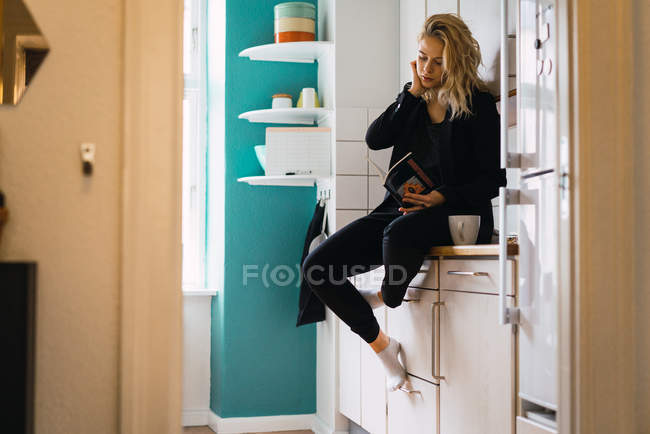 Junge Frau liest Buch, während sie auf einer Tischplatte mit Kaffee sitzt — Stockfoto