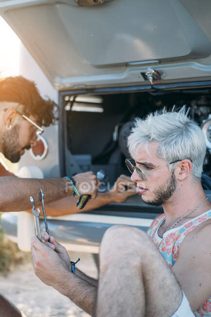 Young male leaning back and relaxing while sitting near friend trying to repair broken motor of car while?during travel in nature — Stock Photo