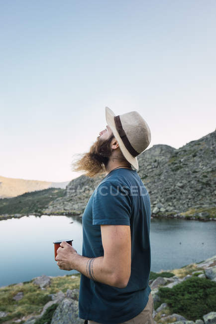 Young relaxed man in hat standing near lake with cup and enjoying view — Stock Photo