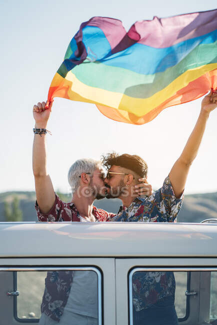 Two handsome men kissing and holding bright LGBT flag while standing inside van with opened roof in countryside on sunny day — Stock Photo