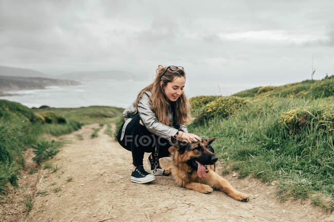 Pretty young female smiling and petting funny German Shepherd while spending time in countryside on cloudy day — Stock Photo
