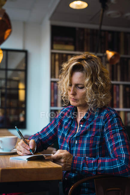 Attractive woman in plaid T-shirt with curly hair sitting in cafe with cup of coffee and writing in notebook with pen — Stock Photo