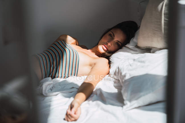 Young pensive woman in striped crop top lying on bed in shadowed bedroom and looking at camera — Stock Photo