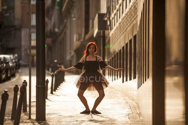 Ballerine à tête rouge avec tutu noir et pointes de ballet rouge dansant dans la rue avec arrière-plan lumineux au coucher du soleil — Photo de stock