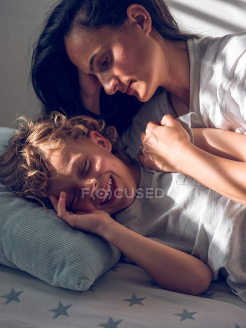 Mulher bonita deitada na cama atrás do menino doce e tocando sua bochecha com cuidado — Fotografia de Stock