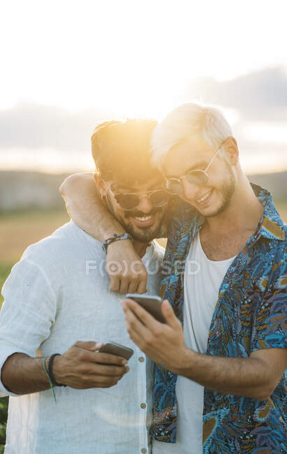 Zwei lächelnde Männer, die sich umarmen und Smartphones schmökern, während sie gemeinsam in der schönen Landschaft stehen — Stockfoto