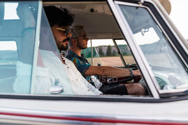 Two handsome men laughing and looking away while embracing each other and riding in retro car during trip — Stock Photo