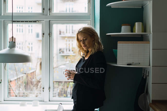 Femme blonde romantique debout avec tasse à la fenêtre à la maison — Photo de stock