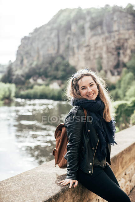 Bella giovane femmina sorridente e guardando la fotocamera mentre si appoggia sul bordo di cemento del ponte su sfondo sfocato di acqua e scogliera — Foto stock