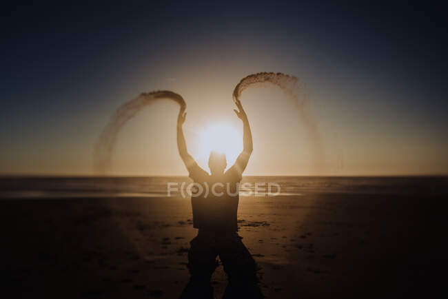 Silhueta de pessoa irreconhecível jogando areia na praia ao pôr do sol de volta iluminado. — Fotografia de Stock