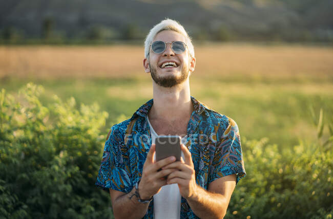 Schöner junger Kerl in stylischem Outfit lacht und surft Smartphone, während er an einem sonnigen Tag vor dem Hintergrund der schönen Natur steht — Stockfoto