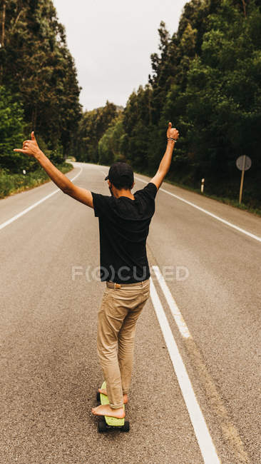 Visão traseira do jovem descalço homem mostrando shaka sinal e andar de skate ao longo da estrada rural — Fotografia de Stock